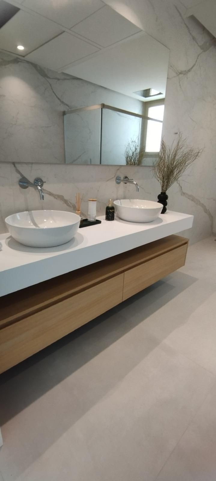 Tocador de baño moderno con dos lavabos blancos sobre encimera, espejo grande y armarios de madera clara.