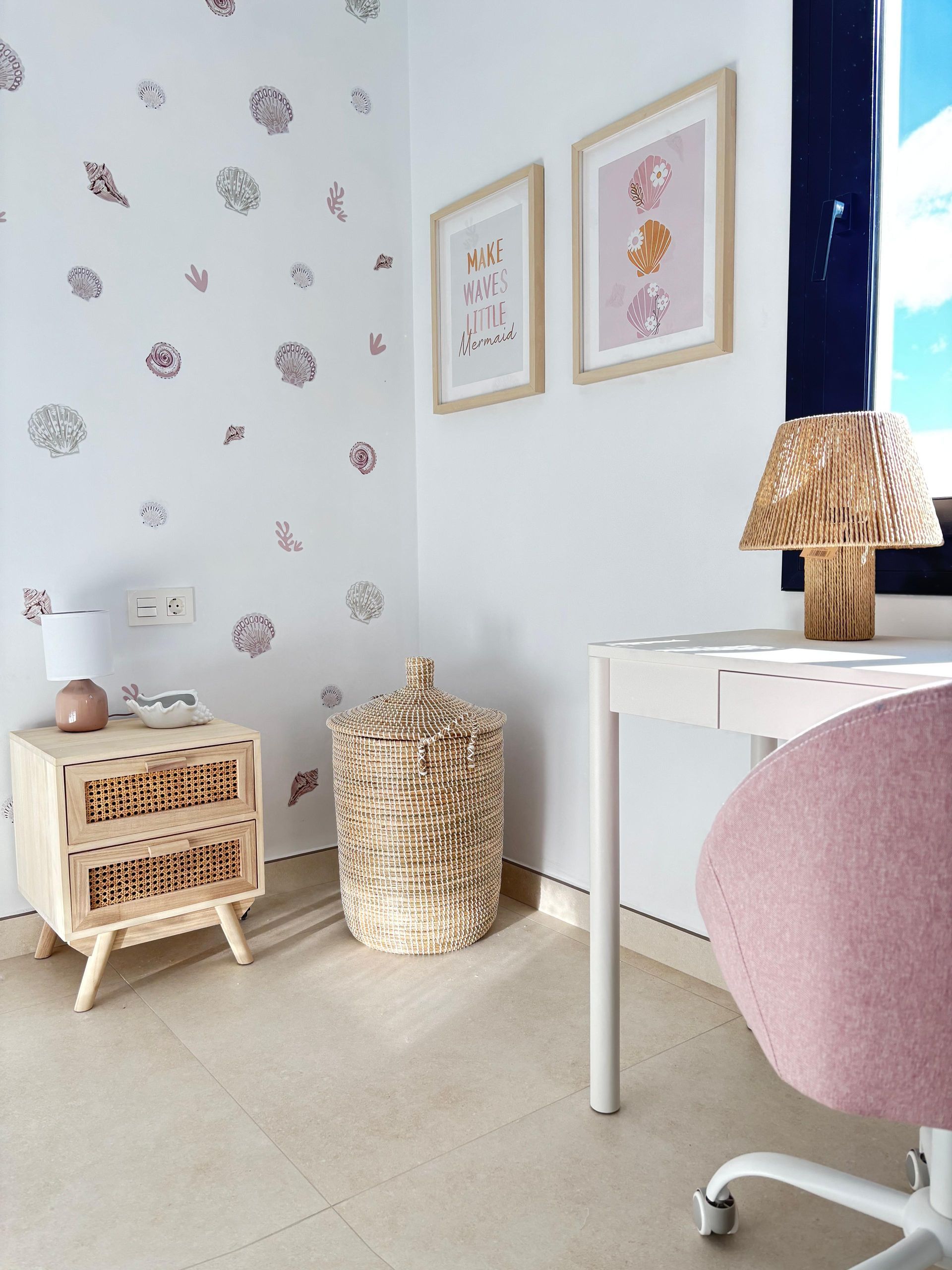 Mesa de comedor de cristal con sillas de mimbre y un jarrón blanco con plantas debajo de dos cuadros enmarcados.