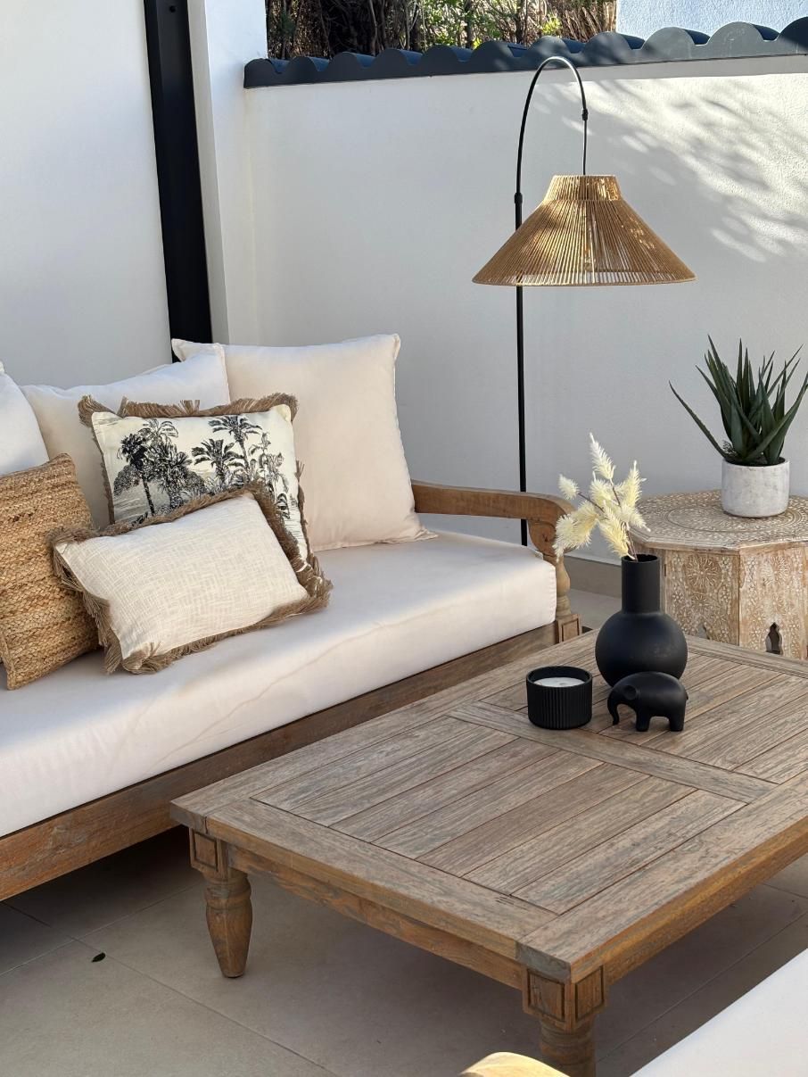 Acogedor patio con sofá blanco, mesa de centro de mimbre, lámpara colgante y planta en maceta en tonos neutros suaves.