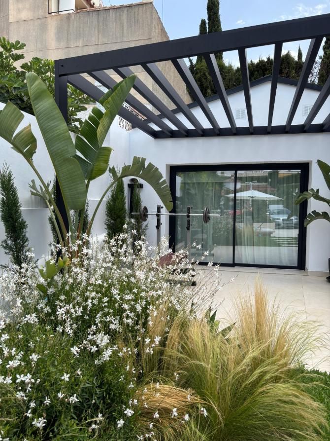 Patio moderno con pérgola negra, puertas de cristal y un exuberante paisaje verde y blanco.