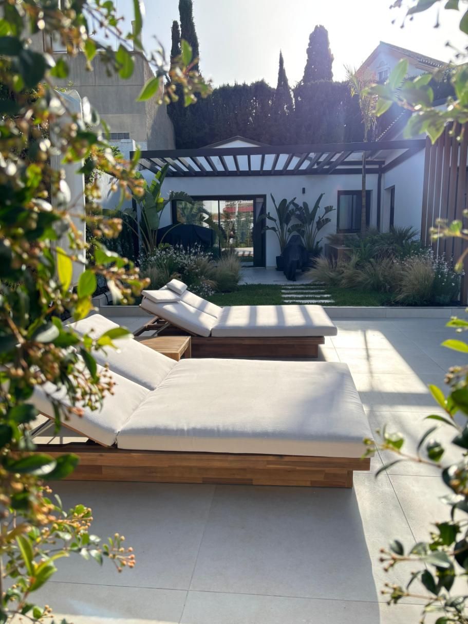 Patio moderno con tumbonas blancas, una cama baja de madera y vegetación en un patio soleado.