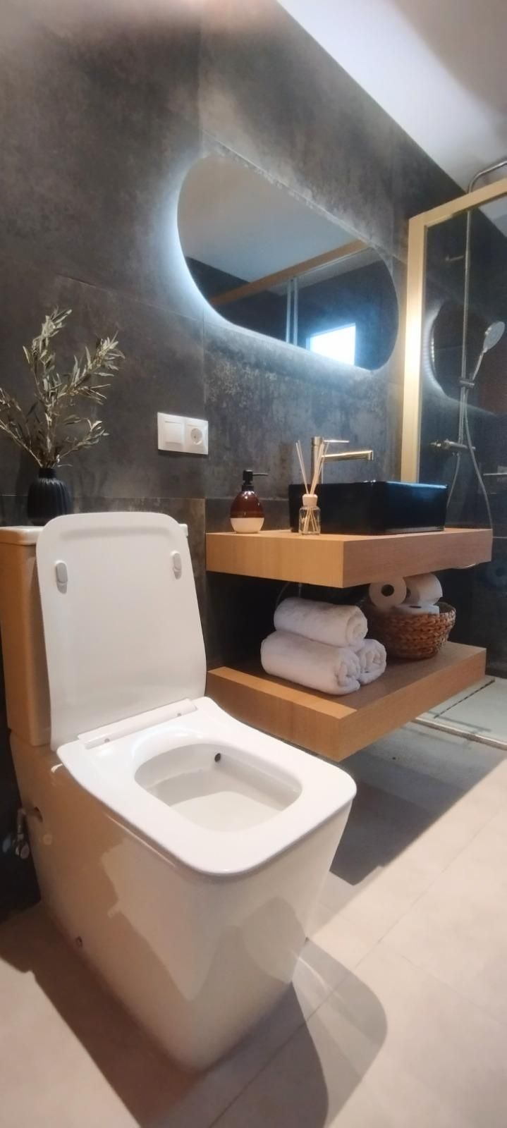 Baño moderno con un inodoro en primer plano y un lavabo de madera con un espejo redondo detrás.