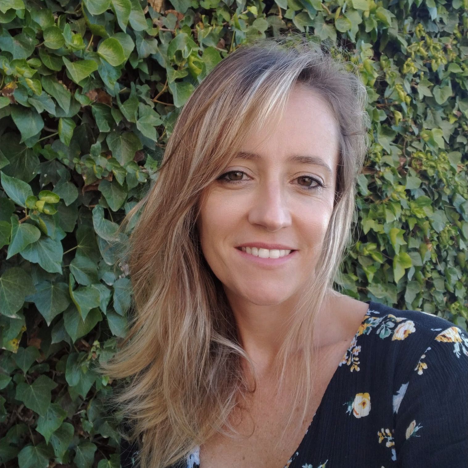 Mujer sonriente con cabello rubio largo y blusa floreada, de pie frente a una pared verde frondosa.