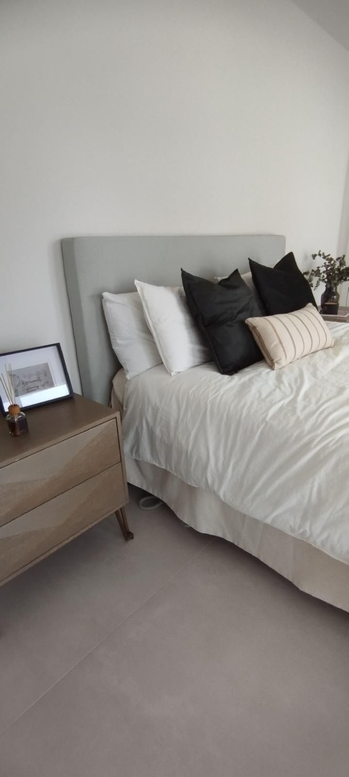 Dormitorio con cama, cabecero gris, ropa de cama blanca, almohadas blancas y negras y una mesita de noche de madera 