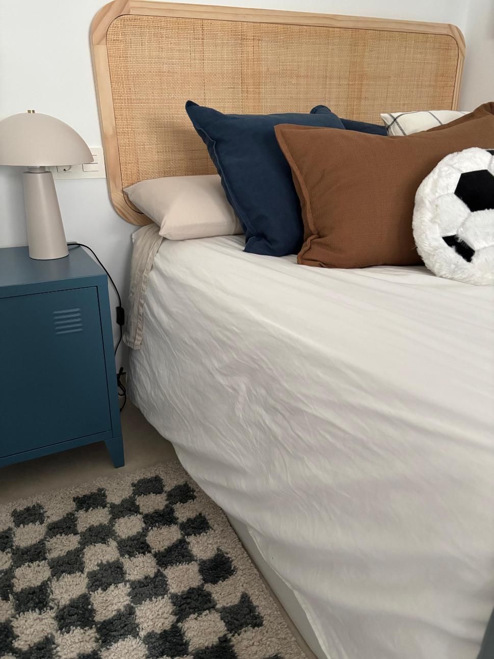 Dormitorio con cabecero tejido, mesita de noche azul, lámpara blanca y almohadas sobre una cama con alfombra a cuadros.