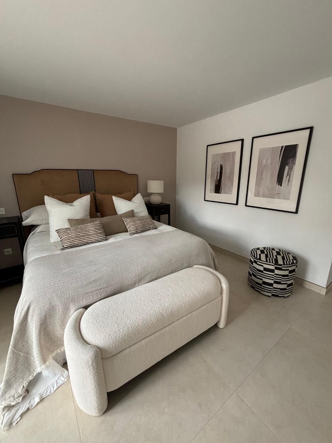 Habitación luminosa con cama, banco beige, cuadro enmarcado en la pared y cesta a rayas en el suelo.