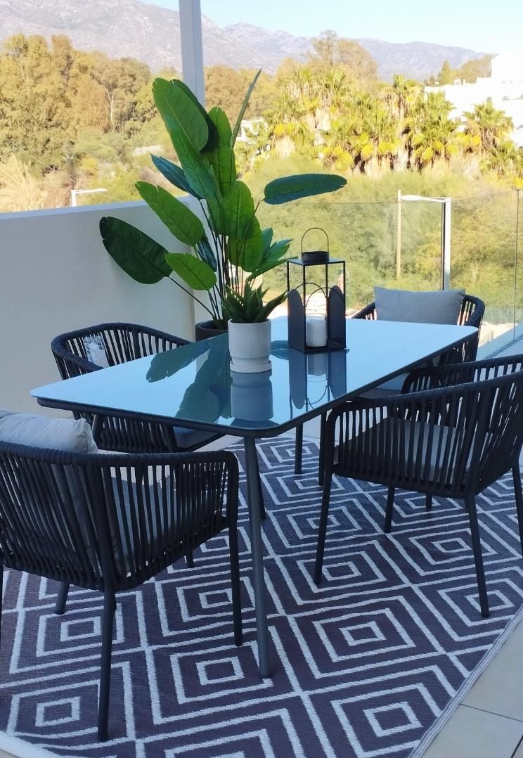 Comedor exterior en el patio con sillas negras, mesa de cristal, alfombra estampada y plantas en macetas.