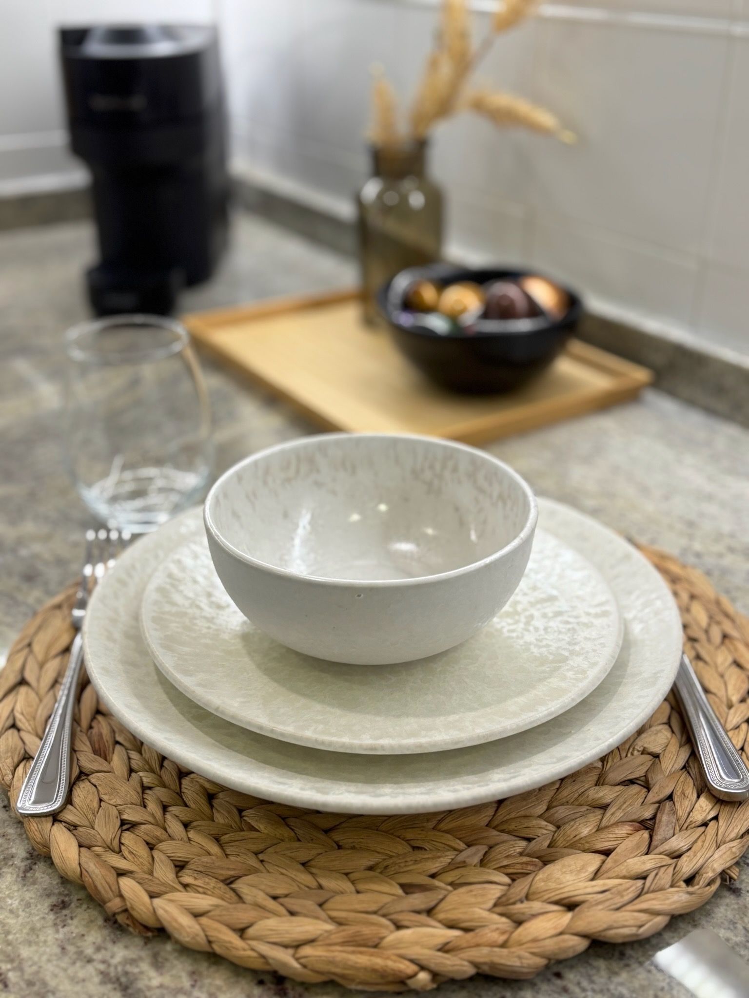 Un cuenco y un plato blancos sobre un mantel individual tejido en una mesa de comedor con una taza negra y un pequeño jarrón al fondo.
