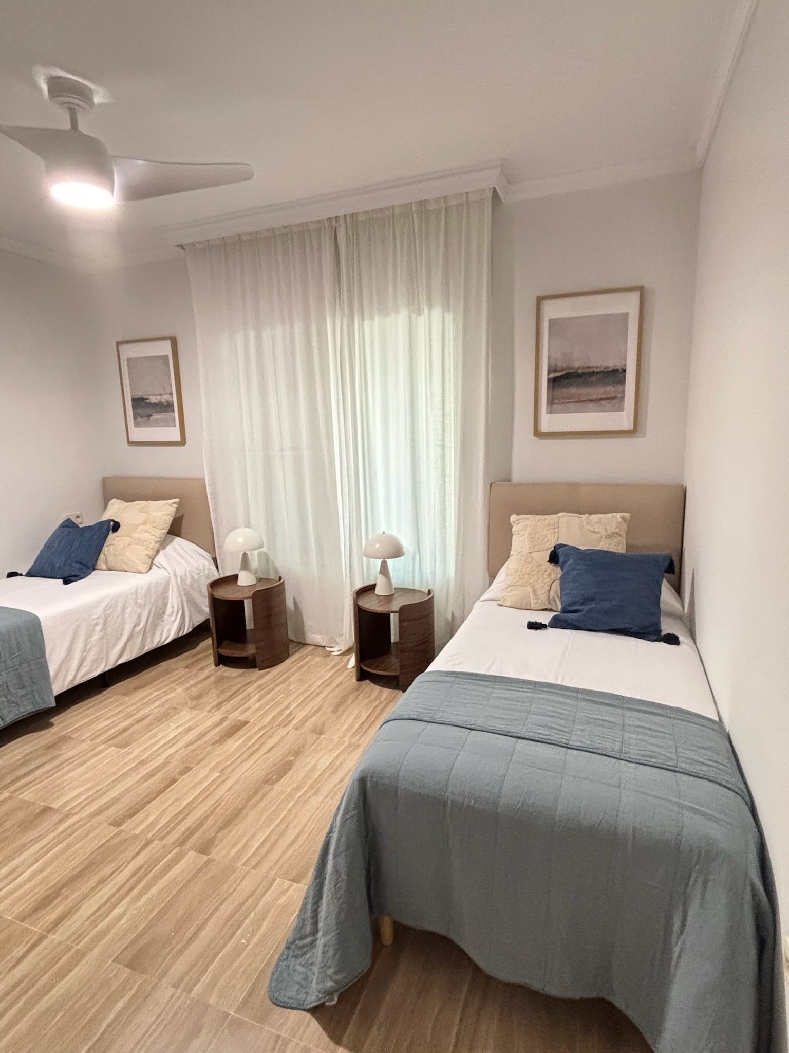 Habitación luminosa con dos camas individuales, mantas azules, cuadros enmarcados y ventilador de techo.