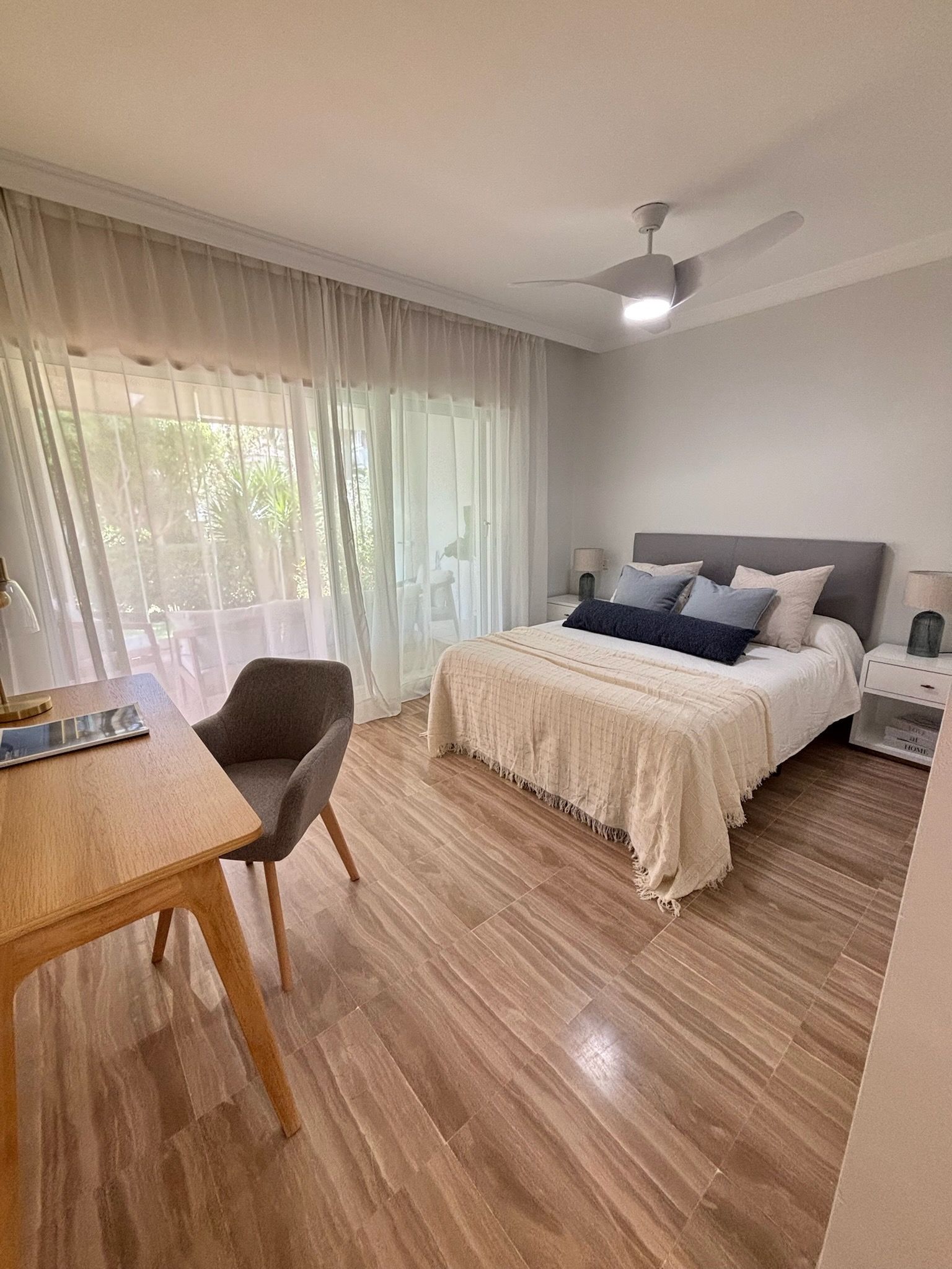 Habitación luminosa con cama, escritorio, silla, suelo de madera y cortinas transparentes junto a una gran ventana.