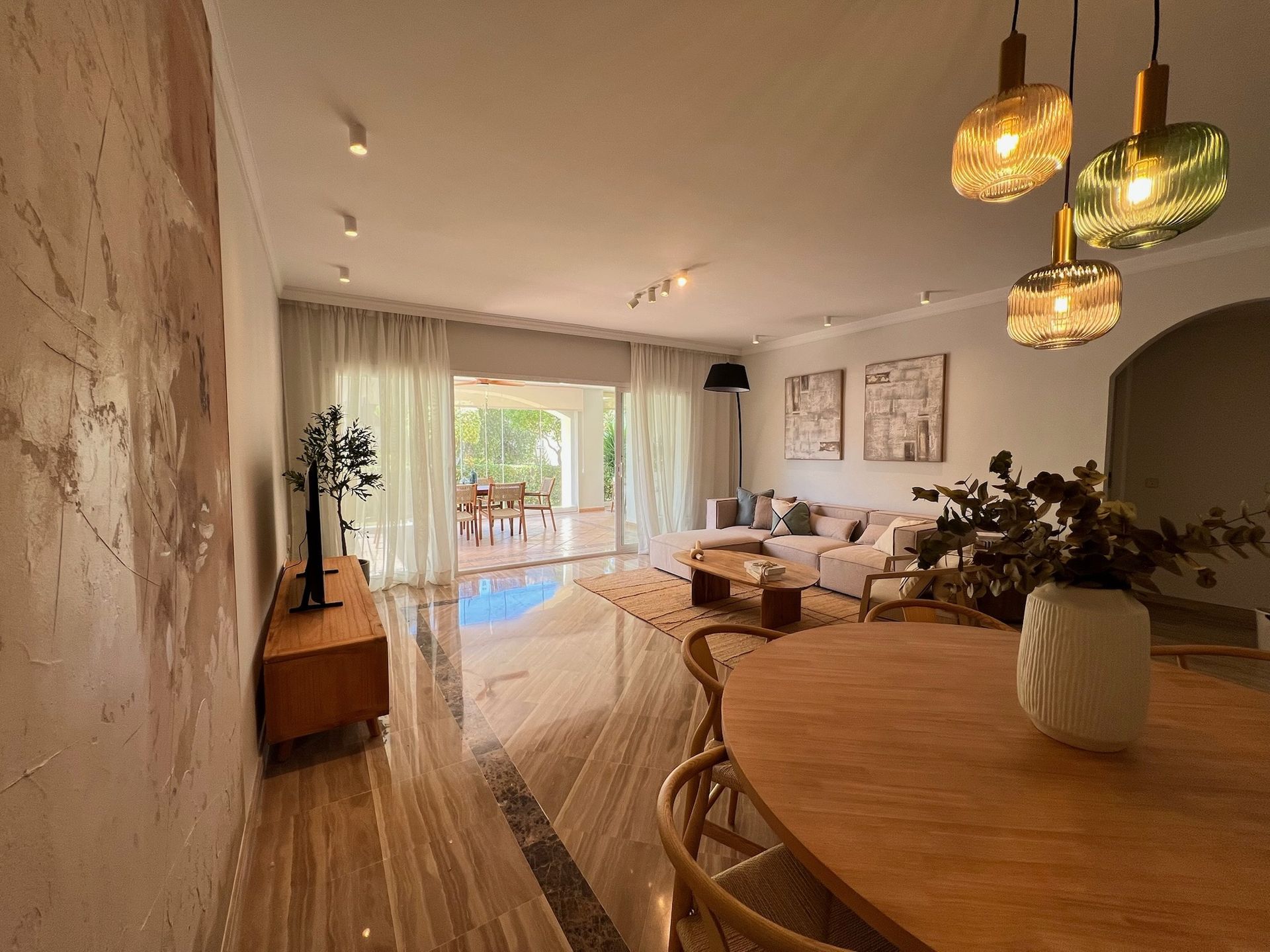 Salón-comedor moderno con mesa de madera, sofá, lámparas colgantes y balcón con vistas.