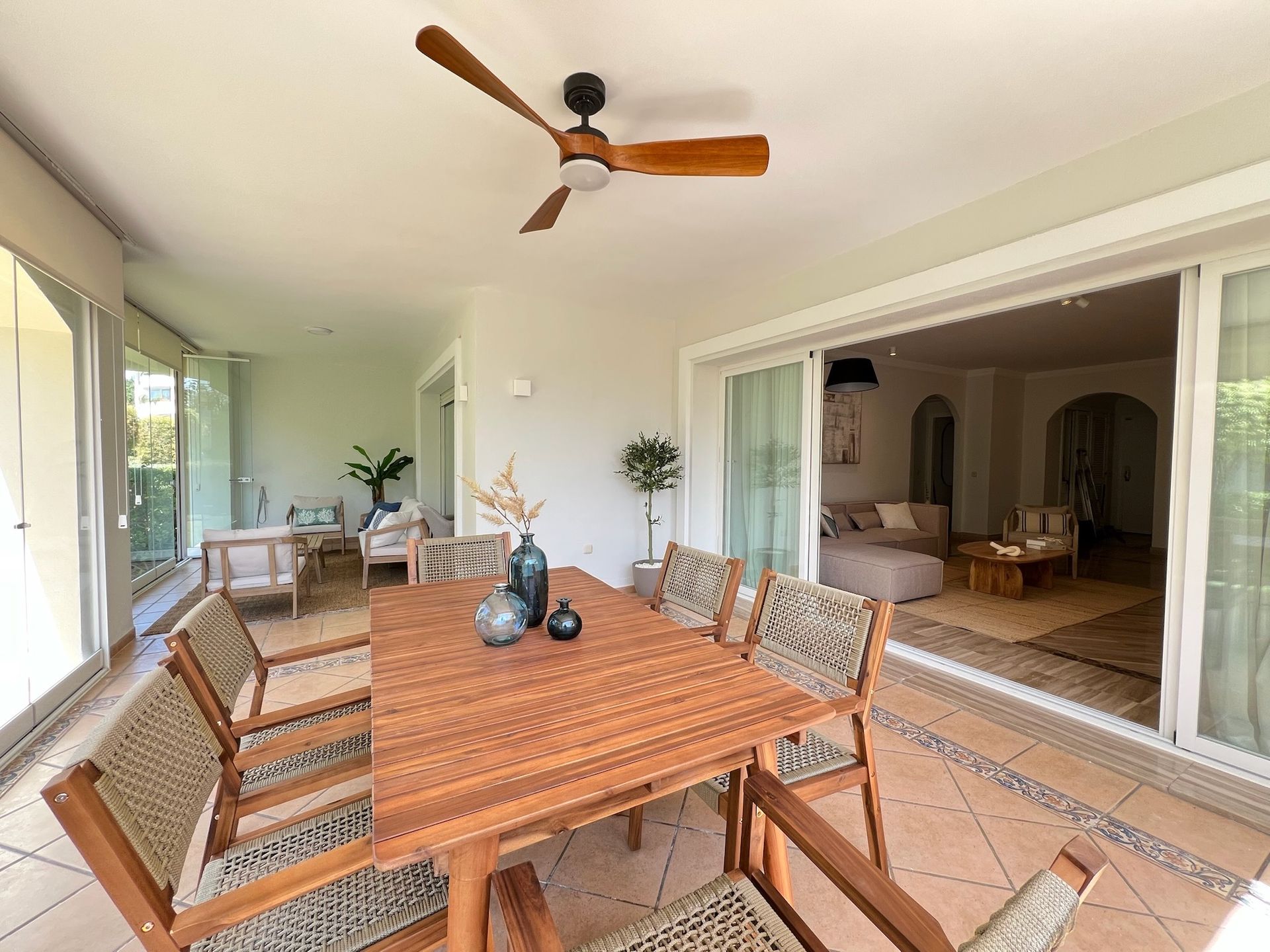 Patio cubierto con mesa de comedor de madera, sillas, ventilador de techo y puertas corredizas que dan a una sala de estar.