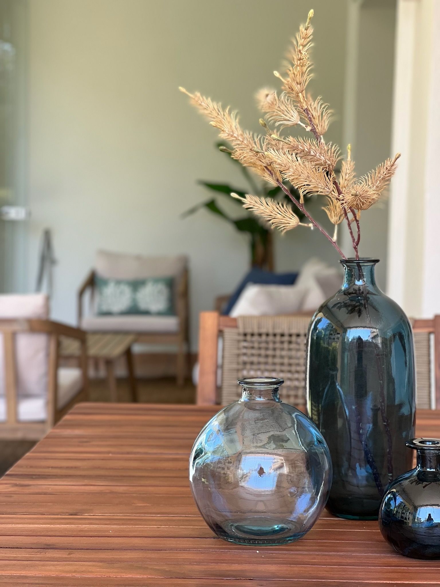 Jarrones de cristal y azul oscuro con flores secas sobre una mesa de madera en una sala de estar con iluminación tenue.