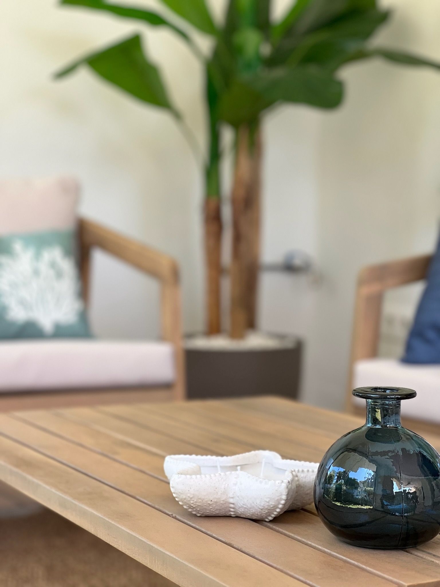 Acogedor patio con mesa de centro de madera, toalla blanca y jarrón de cristal oscuro sobre sillas y plantas difuminadas.
