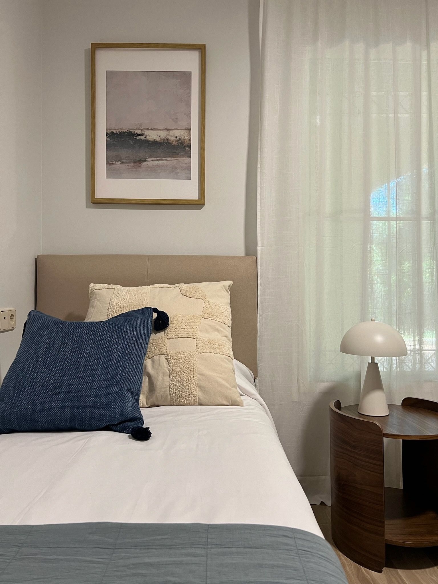 Cama bien hecha con almohadas azules y color crema junto a una mesita de noche redonda con lámpara, un cuadro enmarcado encima y cortinas transparentes.