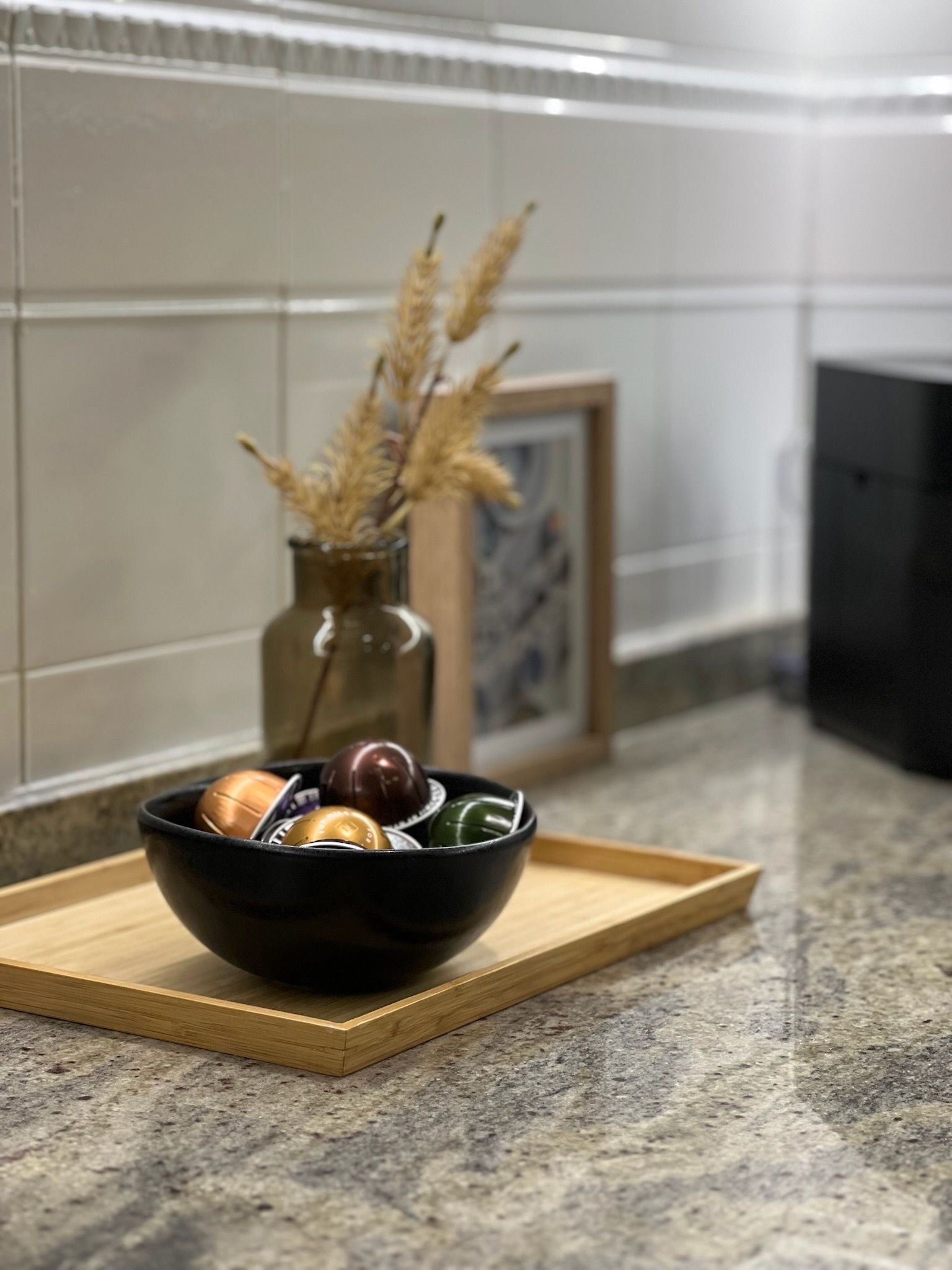Un cuenco negro con frutas de colores sobre una bandeja de madera encima de una encimera de cocina, con un jarrón de trigo seco detrás.