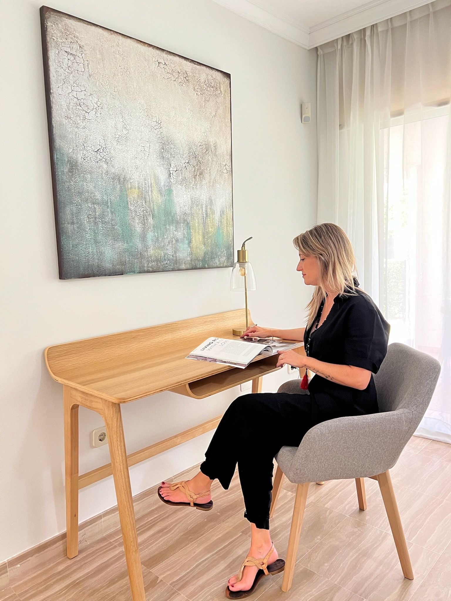 Una mujer sentada en un escritorio en una habitación luminosa, escribiendo en un cuaderno abierto cerca de un gran cuadro abstracto.