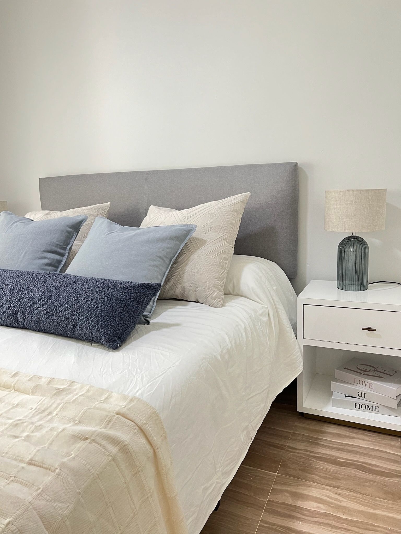 Cama bien hecha con almohadas grises y azules junto a una mesita de noche blanca y una lámpara en un dormitorio luminoso.