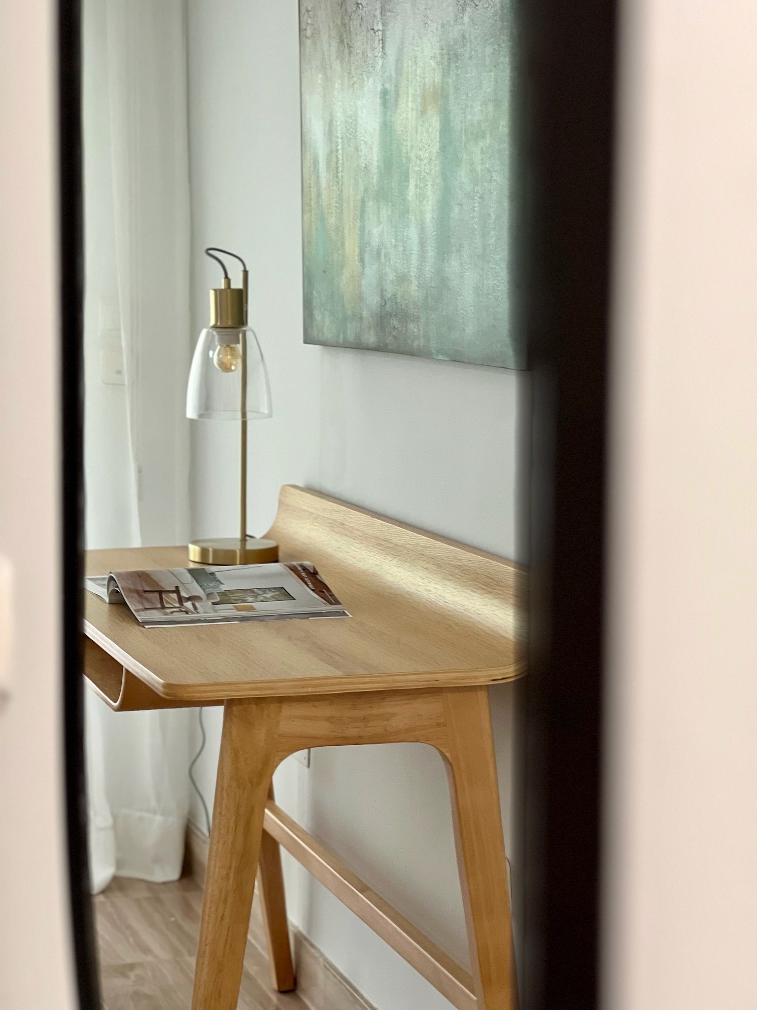 Un escritorio de madera junto a una ventana con una lámpara, revistas y un cuadro abstracto verde en la pared.