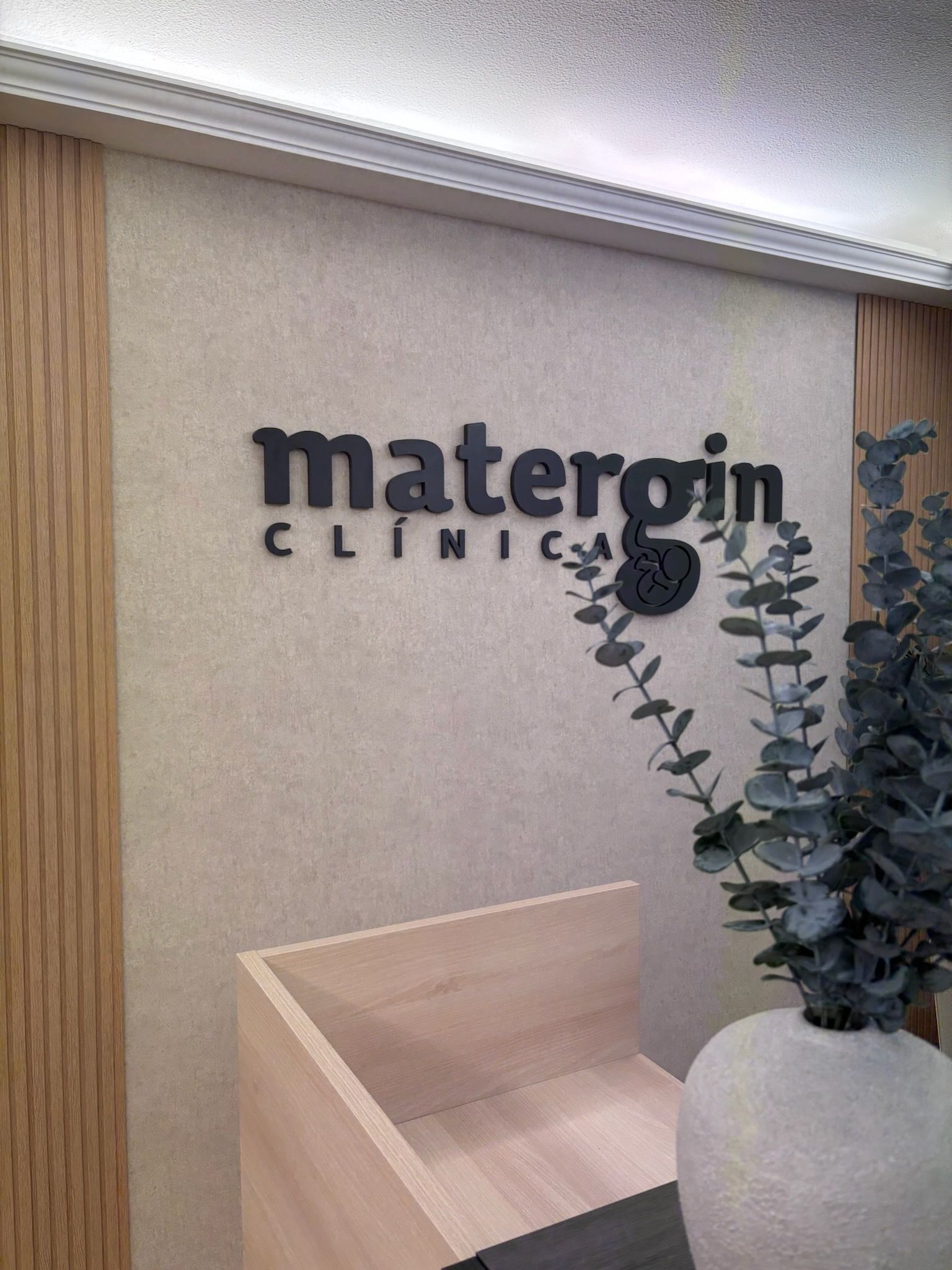 Recepción de la clínica de maternidad con paredes beige, letrero con el logotipo y una planta de eucalipto en maceta.