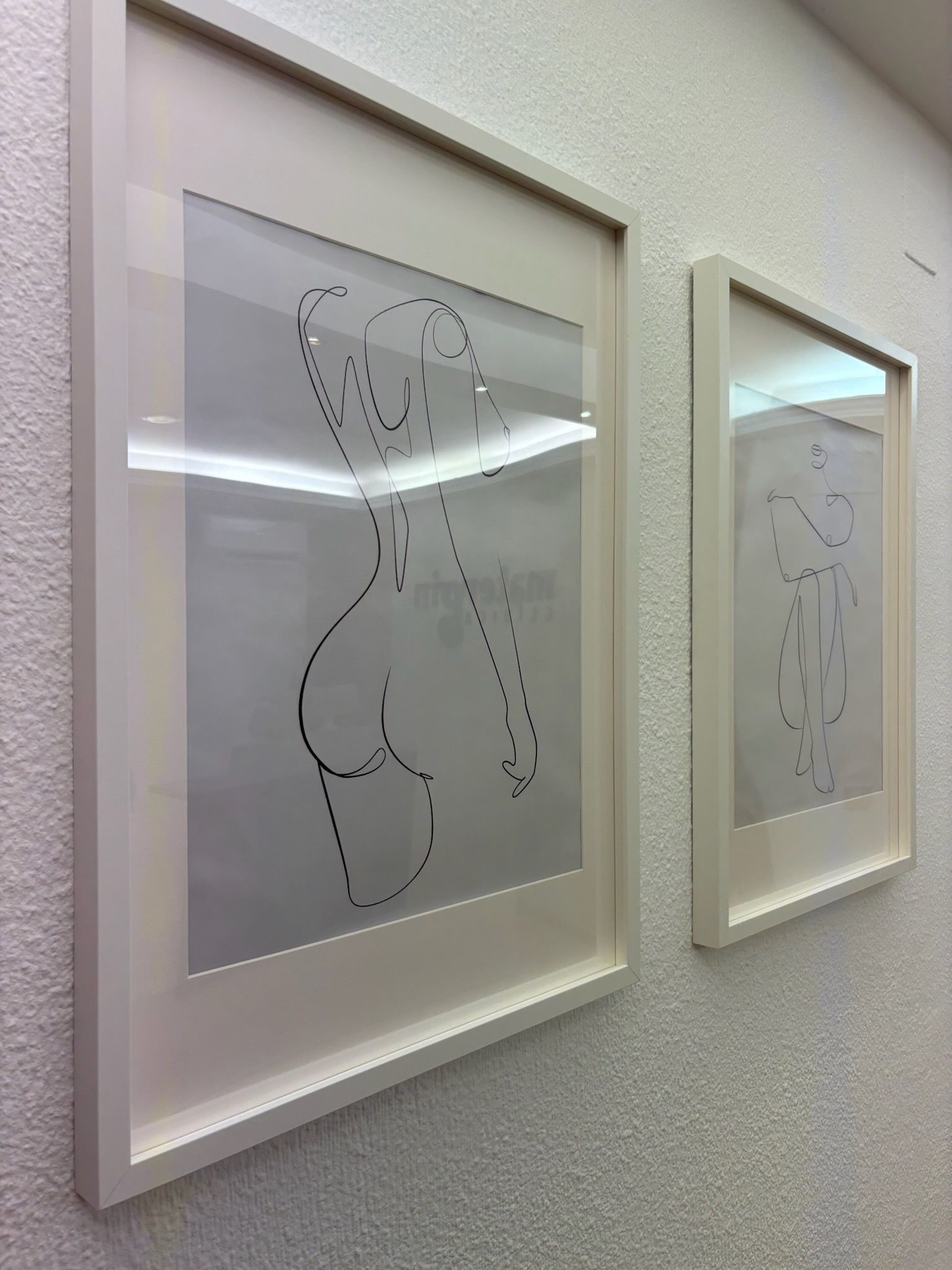 Dos obras de arte enmarcadas en vidrio esmerilado sobre una pared texturizada, cada una con un dibujo de un torso dibujado con líneas punteadas.