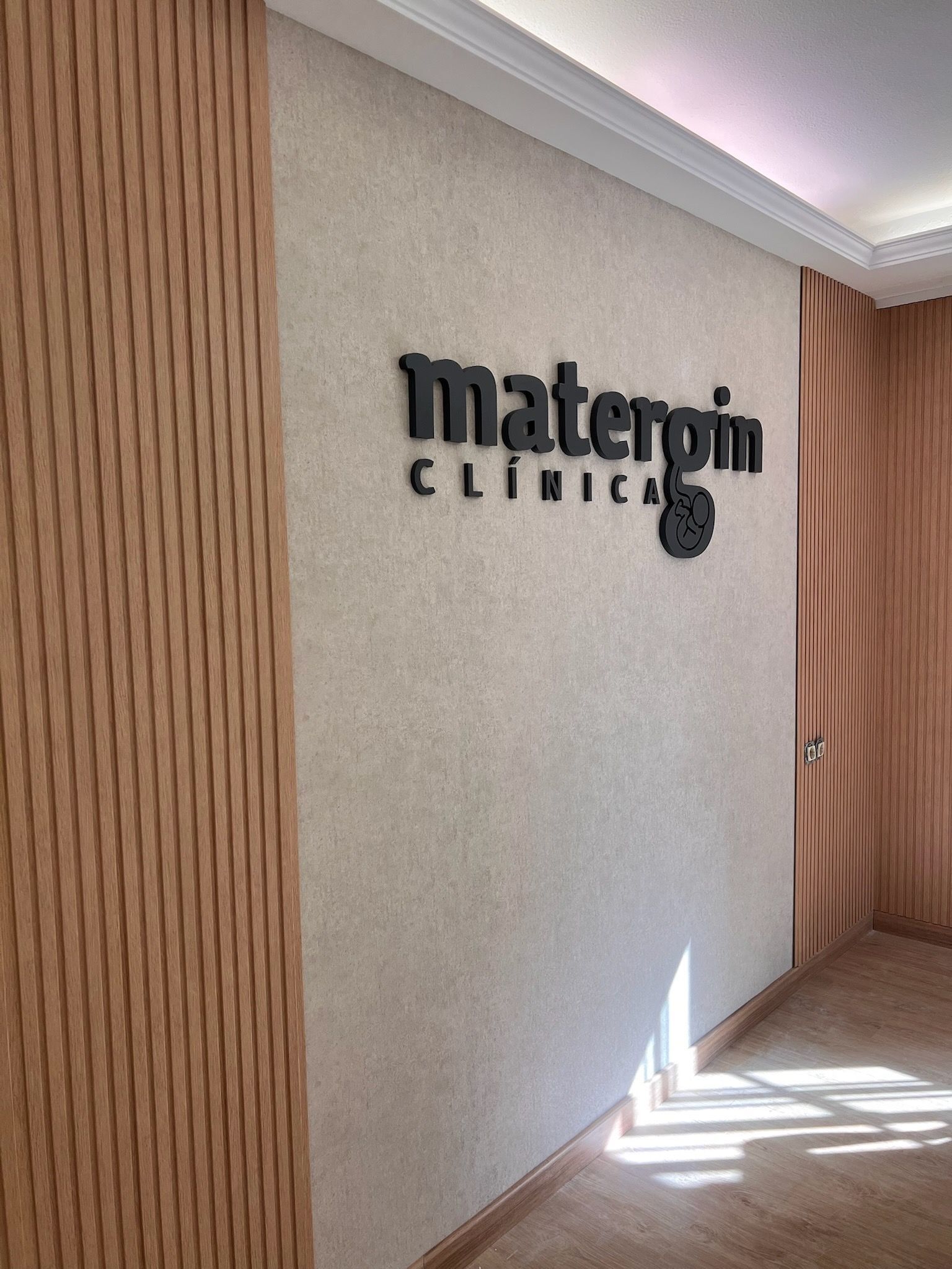 Logotipo de la clínica en una pared texturizada en un pasillo luminoso con paneles de madera y luz solar en el suelo.