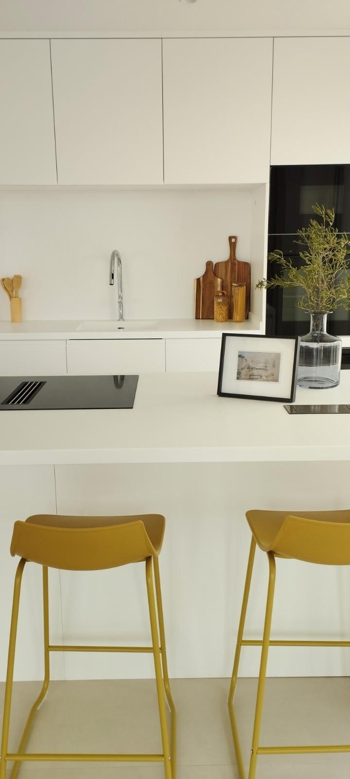 Cocina moderna blanca con una isla amarilla, dos taburetes color mostaza y un jarrón de flores.