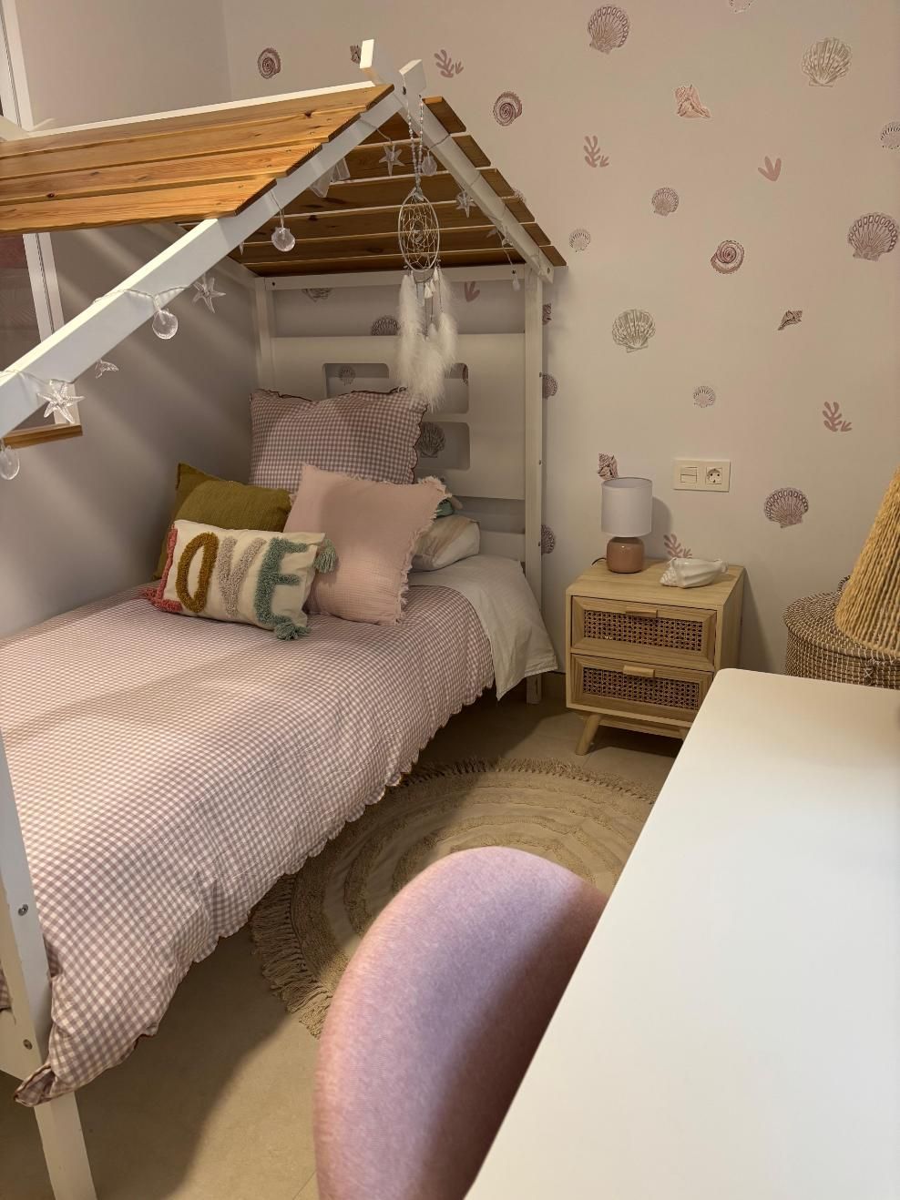 Habitación infantil con cama con forma de casita, ropa de cama rosa, escritorio y mesita de noche tejida.