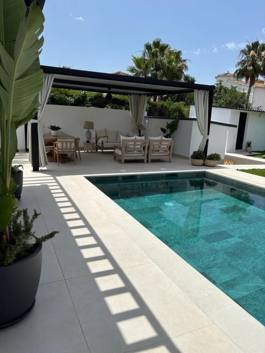 Patio junto a la piscina con pérgola, asientos de mimbre y una piscina turquesa bañada por el sol.