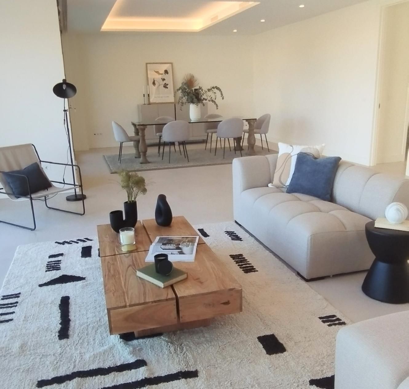 Luminoso salón moderno con sofá blanco, mesa de centro de madera, alfombra y zona de comedor al fondo.