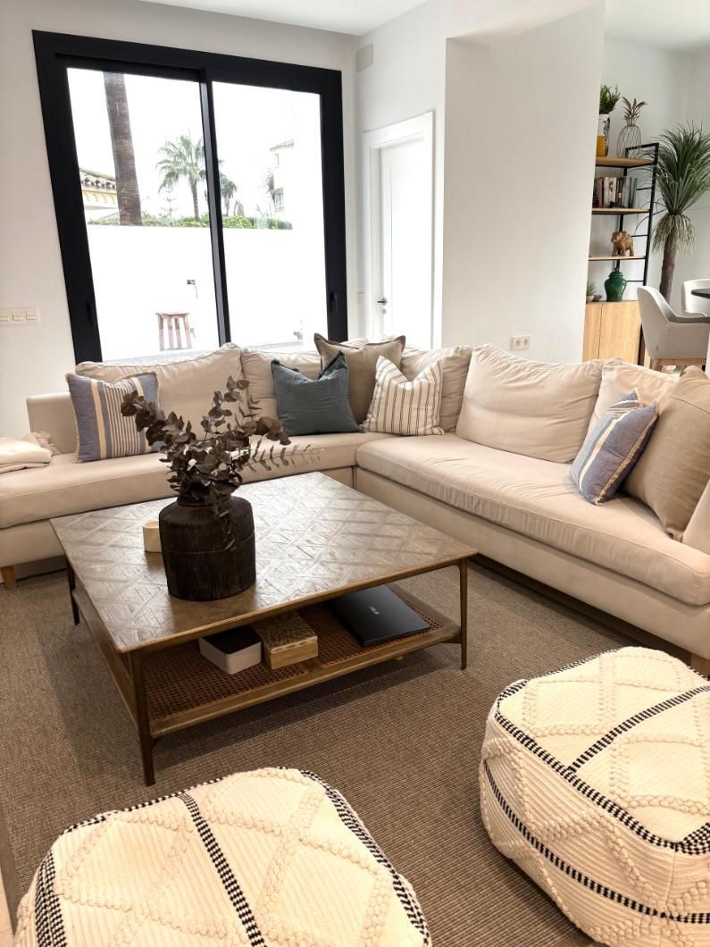 Luminoso salón con sofá modular beige, mesa de centro y gran ventanal con palmeras en el exterior.