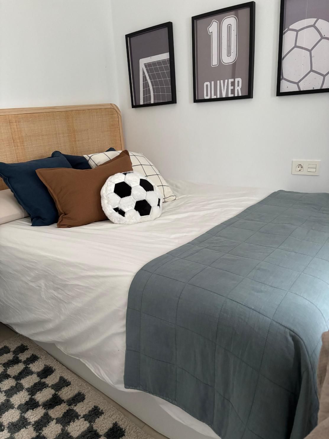 Habitación acogedora con cama blanca, manta azul, almohada con forma de balón de fútbol y cuadro enmarcado sobre el cabecero.