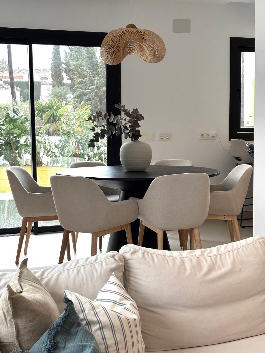 Comedor luminoso con mesa redonda negra, sillas beige, lámpara colgante tejida y sofá en primer plano.