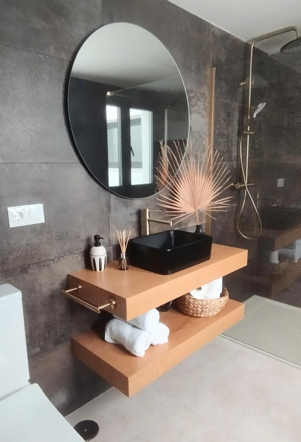 Tocador de baño moderno con espejo redondo, lavabo negro, estantes de madera y elementos decorativos.