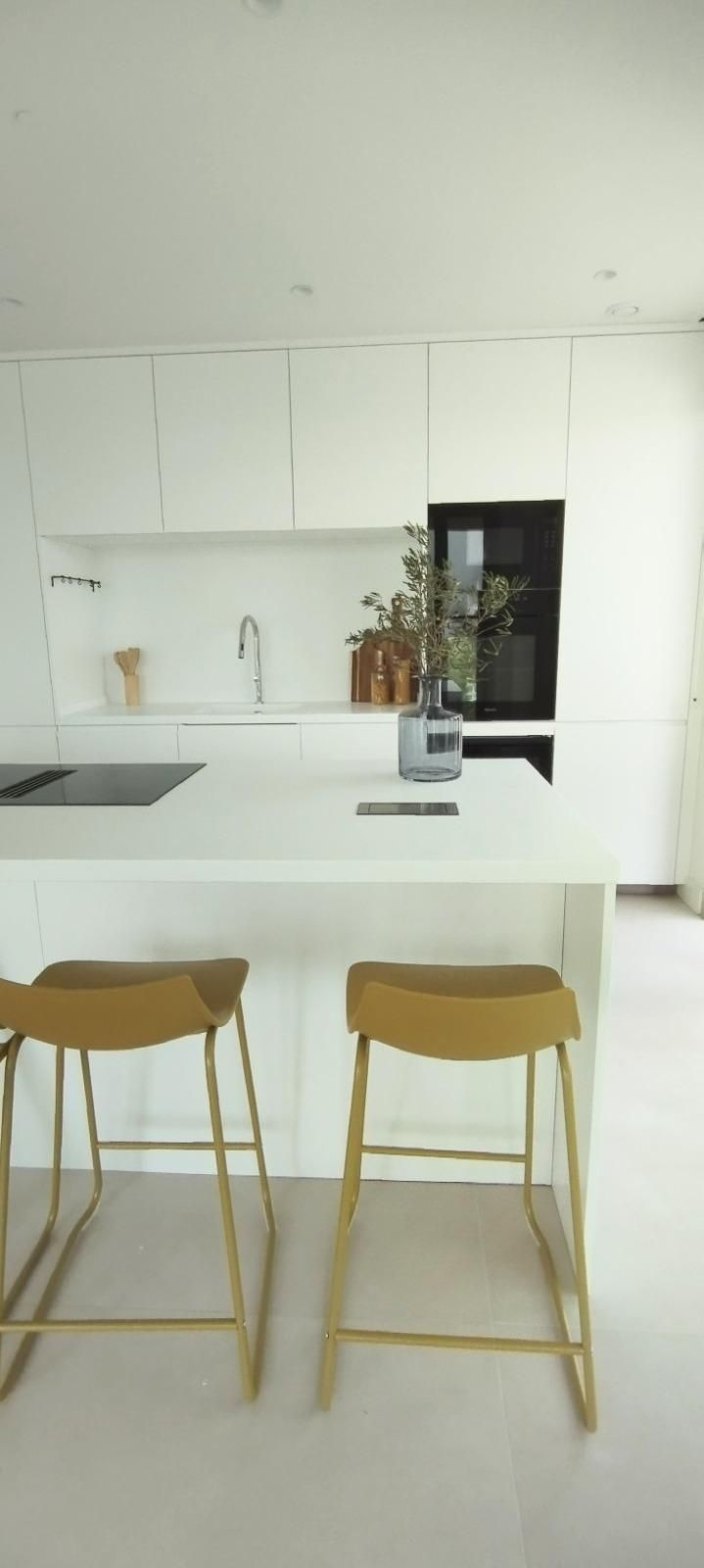 Cocina minimalista blanca con dos taburetes dorados y un frigorífico negro.