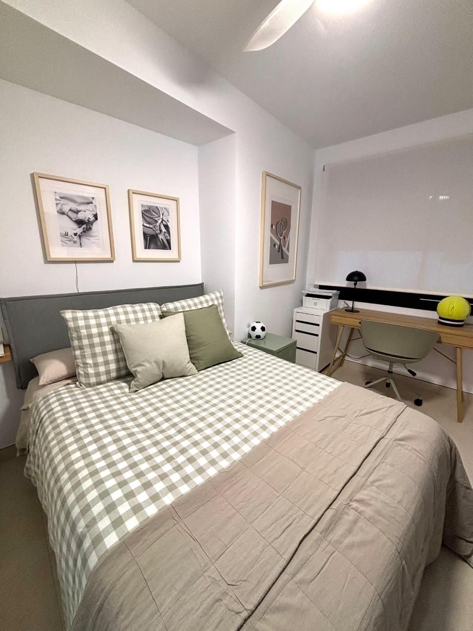 Habitación pequeña con una cama, ropa de cama a cuadros, cuadros enmarcados en la pared y un escritorio junto a la ventana.