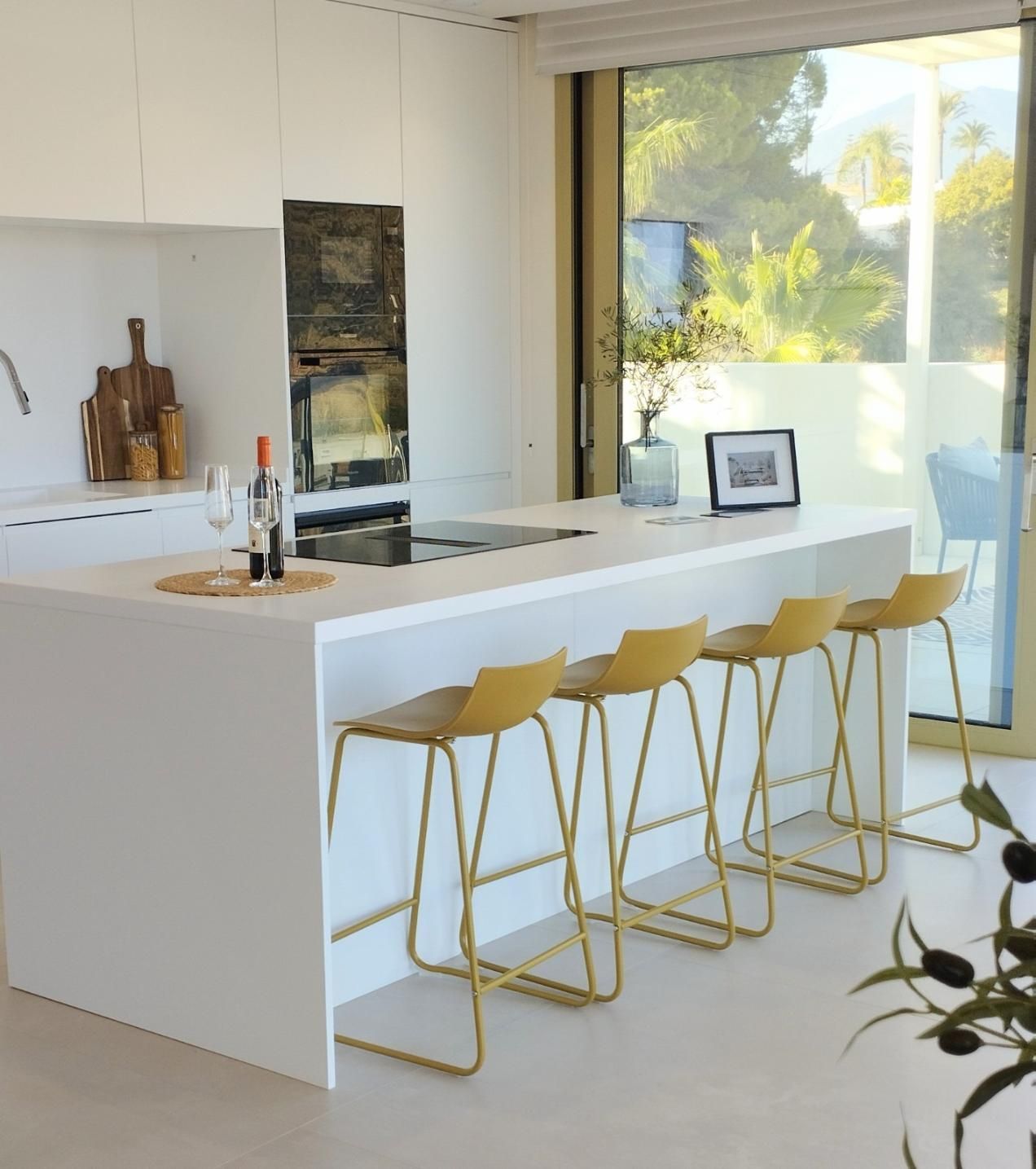 Cocina moderna blanca con taburetes dorados y una gran ventana que deja entrar la luz del sol.