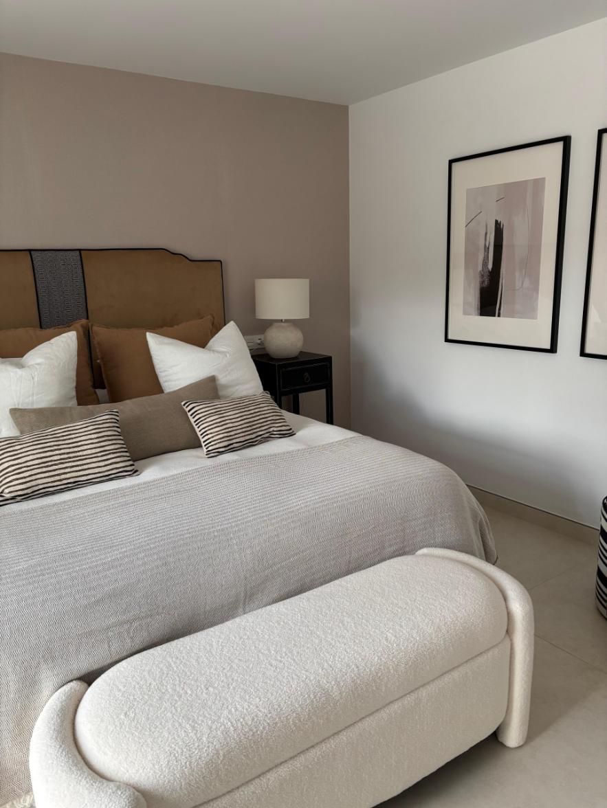 Dormitorio moderno con cama, otomana blanca, lámpara de noche y cuadro enmarcado.