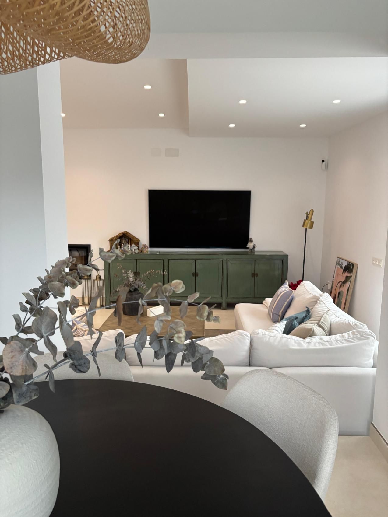 Sala de estar luminosa y moderna con sofás blancos, televisor sobre consola verde y decoración floral seca en primer plano.