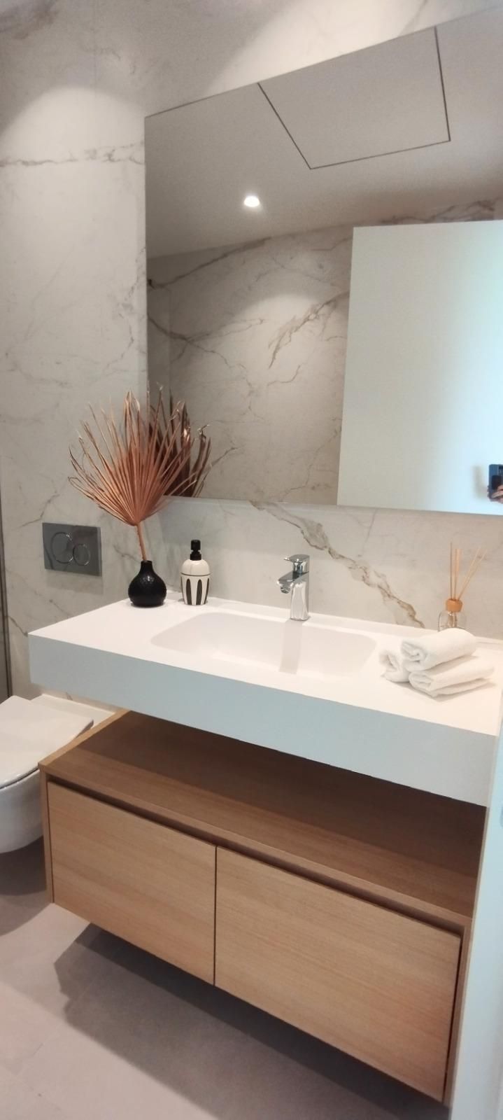 Tocador de baño moderno con lavabo blanco, espejo grande y cajones de madera clara bajo una pared con aspecto de mármol.