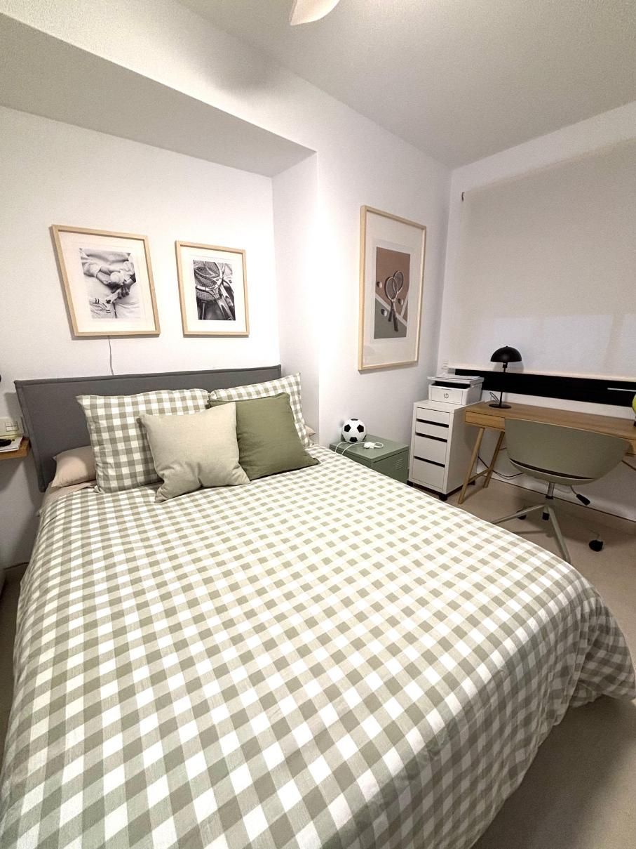 Habitación pequeña con una cama, ropa de cama a cuadros, cuadros enmarcados en la pared y un escritorio junto a la ventana.