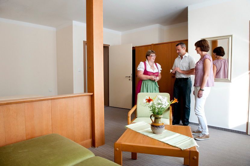 Drei Personen unterhalten sich in einem Hotelzimmer, eine davon in traditioneller Kleidung. Im Vordergrund stehen ein Tisch mit Blumen und ein kleines Sofa.