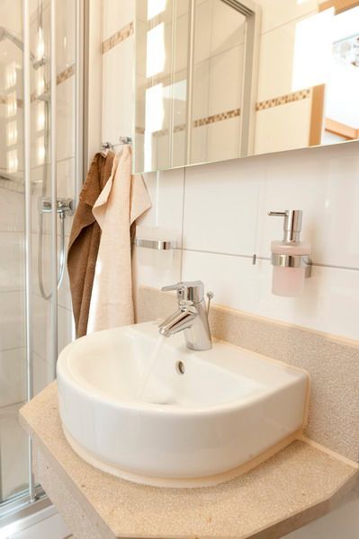 Weißes Waschbecken mit Chromhahn, hellbraune Arbeitsplatte, hängende Handtücher und Dusche dahinter.