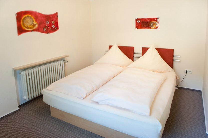 Doppelbett in einem Zimmer mit weißen Wänden, roten Kopfteilen und abstrakter Kunst, Heizkörper an der Wand.