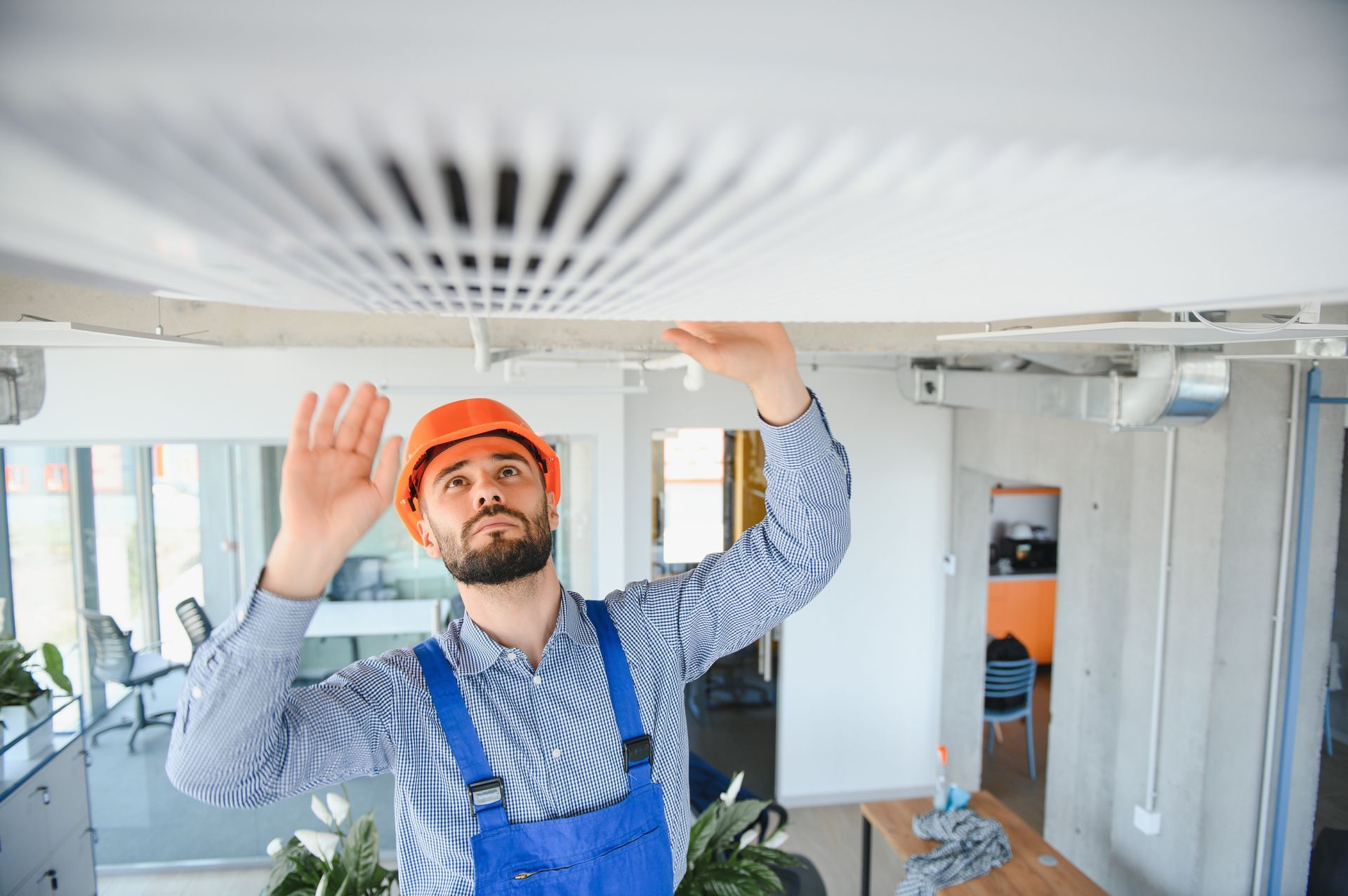 Hombre con mono y casco inspeccionando un respiradero del techo en una oficina.