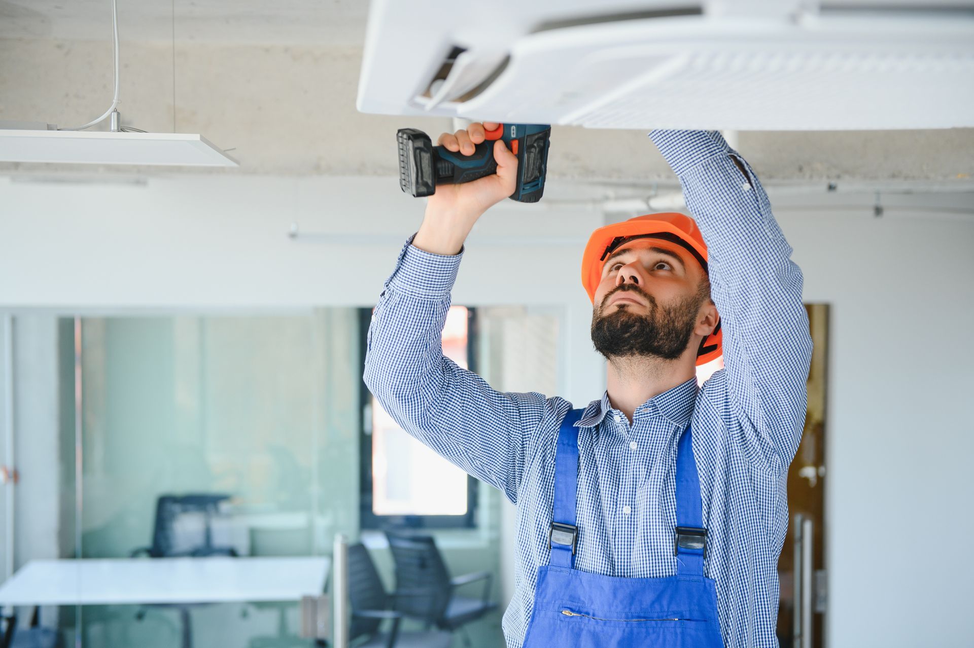 Un trabajador con un mono azul y un casco naranja instala una unidad de techo con un taladro eléctrico.