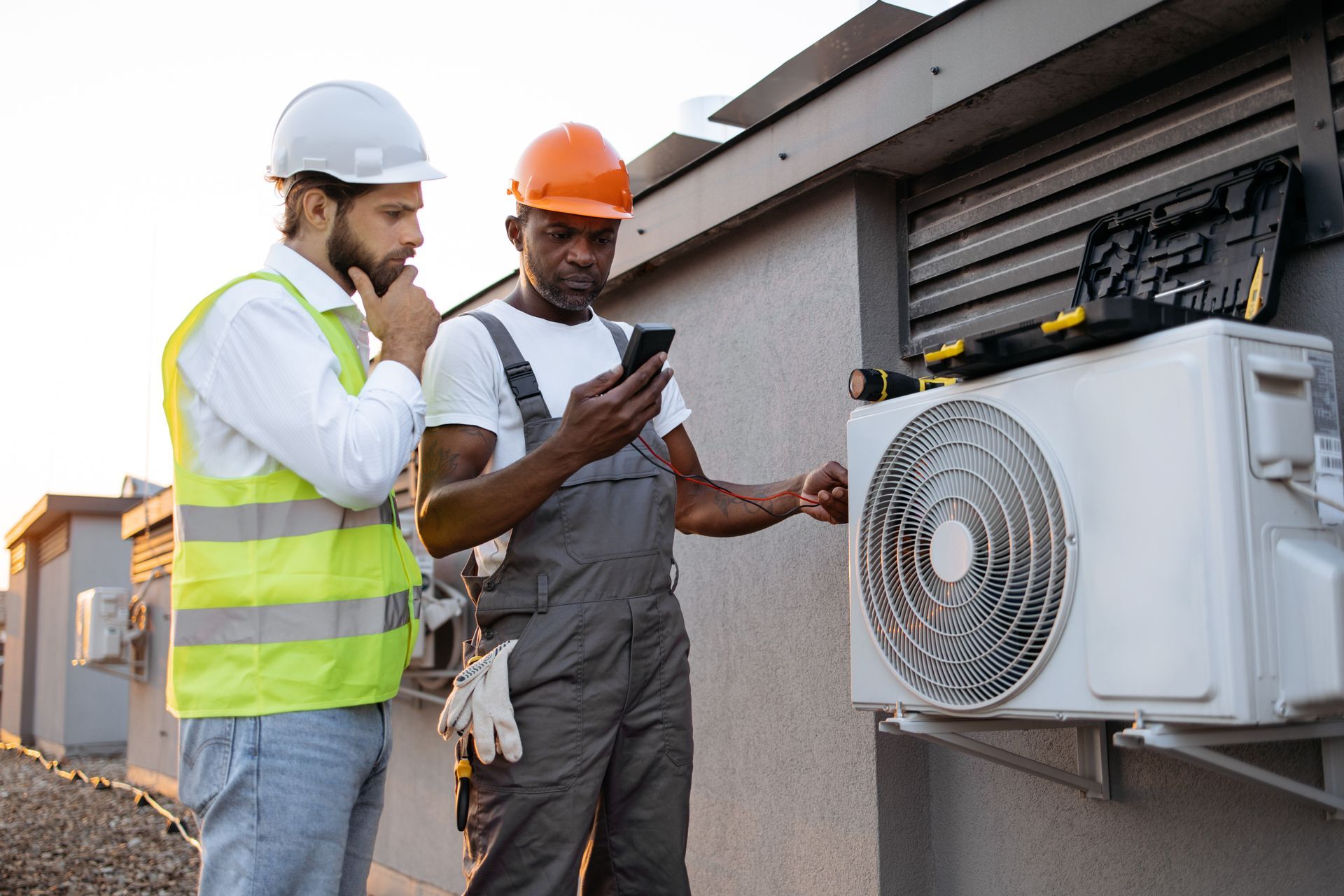 Dos técnicos de HVAC inspeccionan una unidad de aire acondicionado en el exterior, uno mirando un teléfono, el otro pensando.