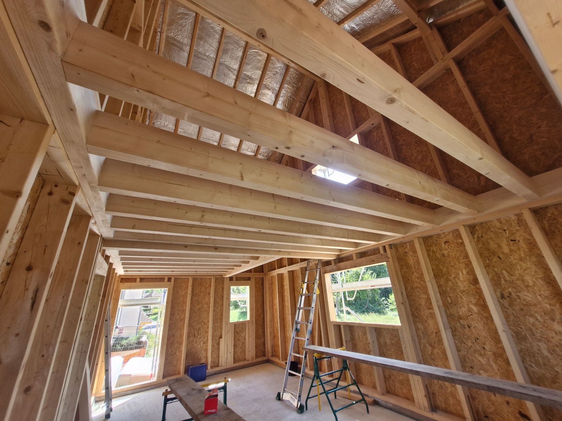 Intérieur d'un bâtiment à ossature en bois en construction avec poutres apparentes, fenêtres et échelle.