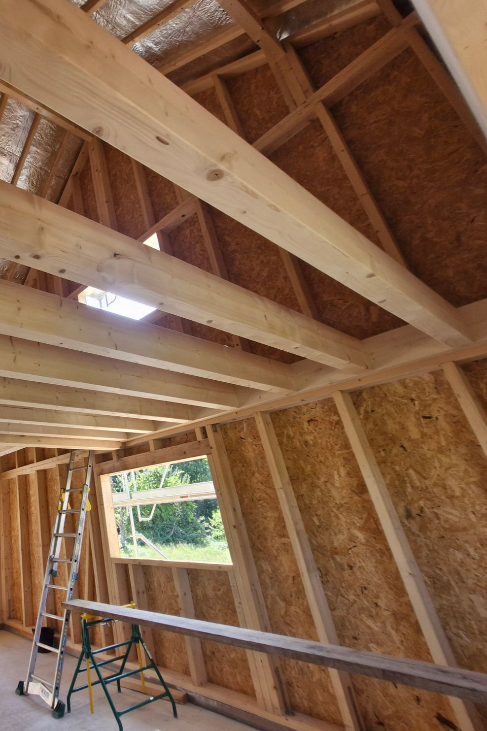 Vue intérieure d'un bâtiment en bois partiellement construit, avec poutres, murs et fenêtre apparents. Une échelle est visible.