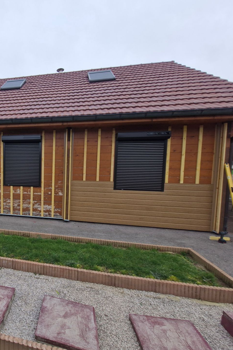 Bâtiment en bois avec toit brun, volets noirs et bardage partiel. Espace gazonné devant.